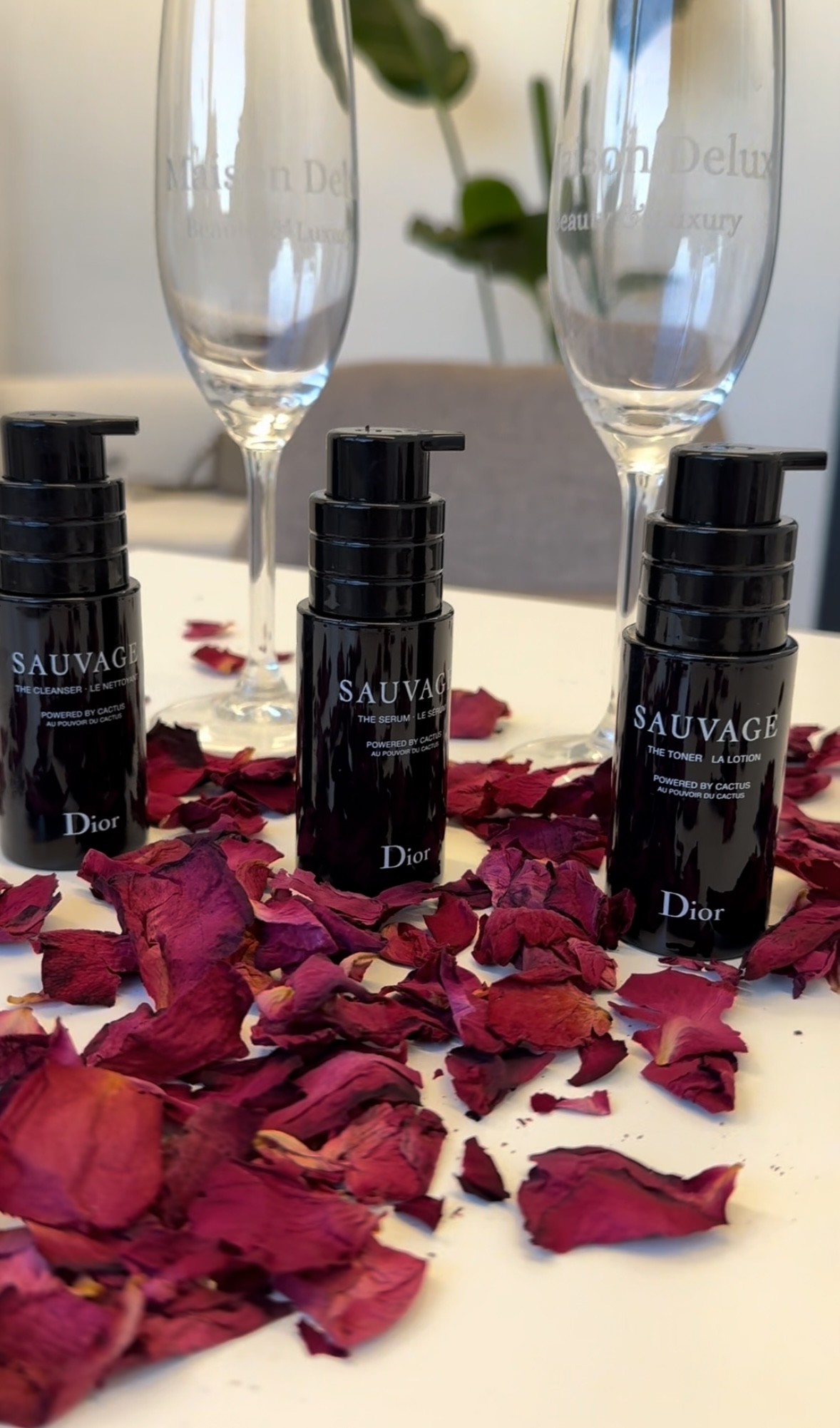 Drie flessen Dior Sauvage met champagneglazen waarin Maison Delux gegraveerd staat, tussen rozenblaadjes